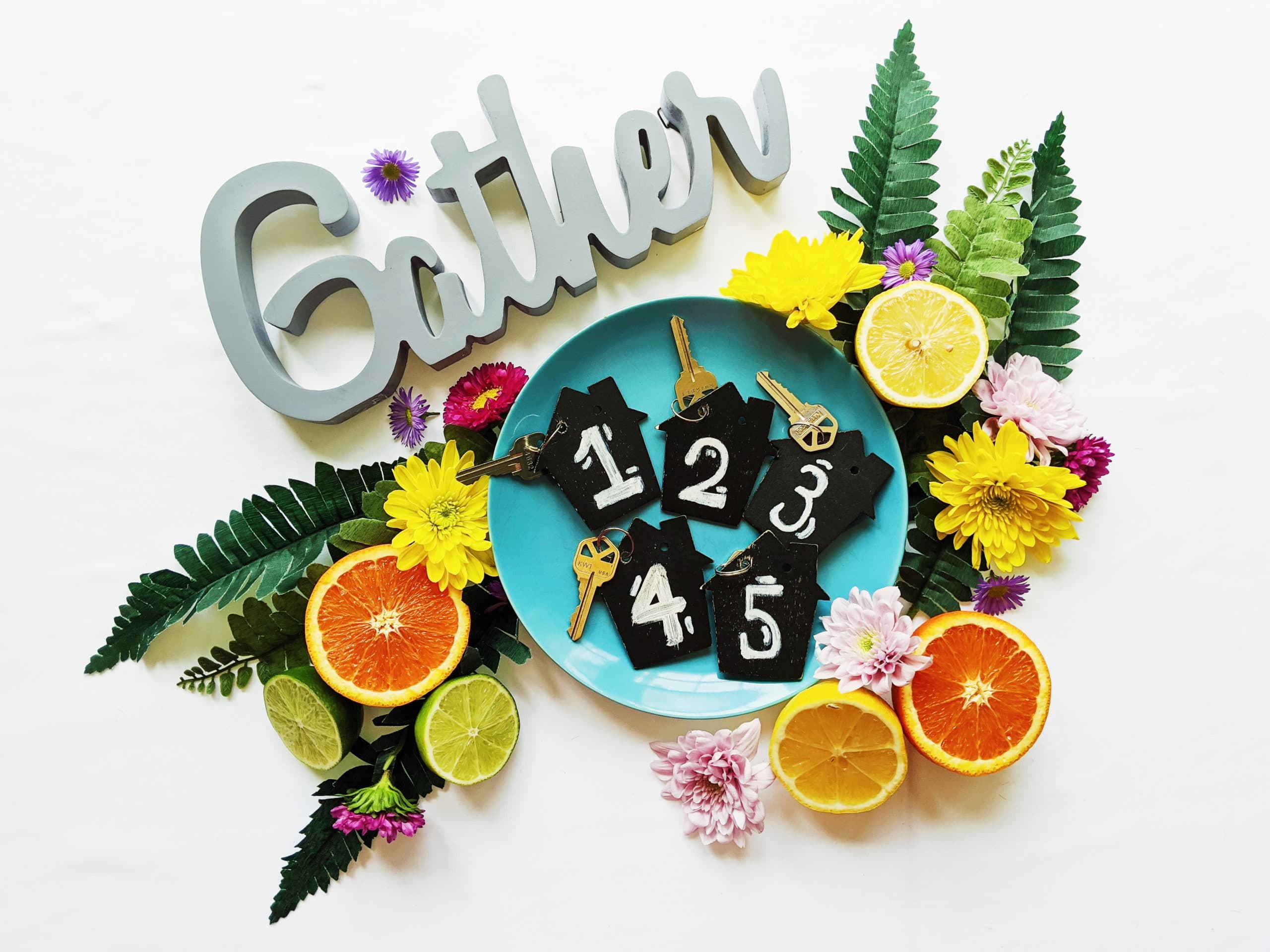 A blue plate with numbered tags and keys, surrounded by colorful flowers and citrus slices, beside a decorative "Gather" sign.