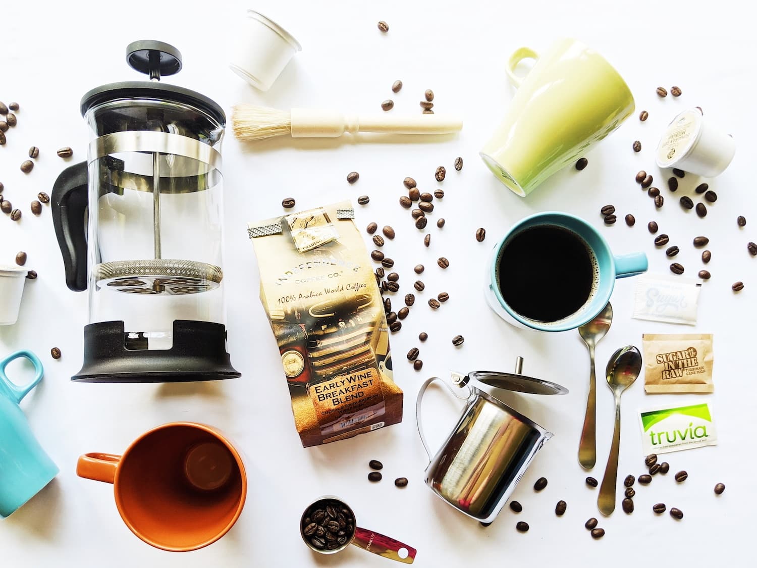 An assortment of coffee-making tools and ingredients, including a French press, coffee mugs, beans, sweeteners, and a measuring spoon arranged on a white surface.