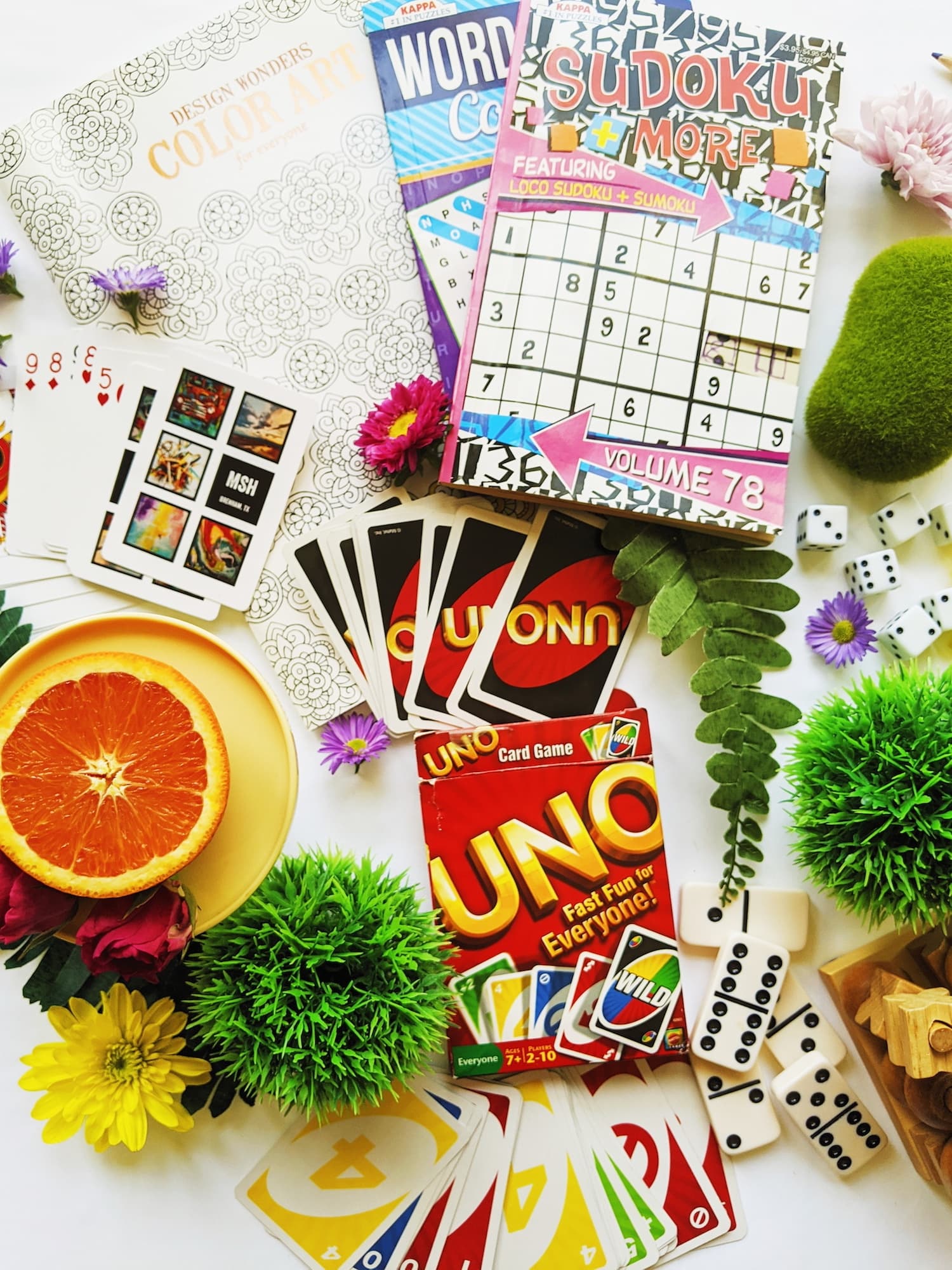 A colorful arrangement of games, including UNO cards, Sudoku and word puzzle books, surrounded by fruits and flowers.