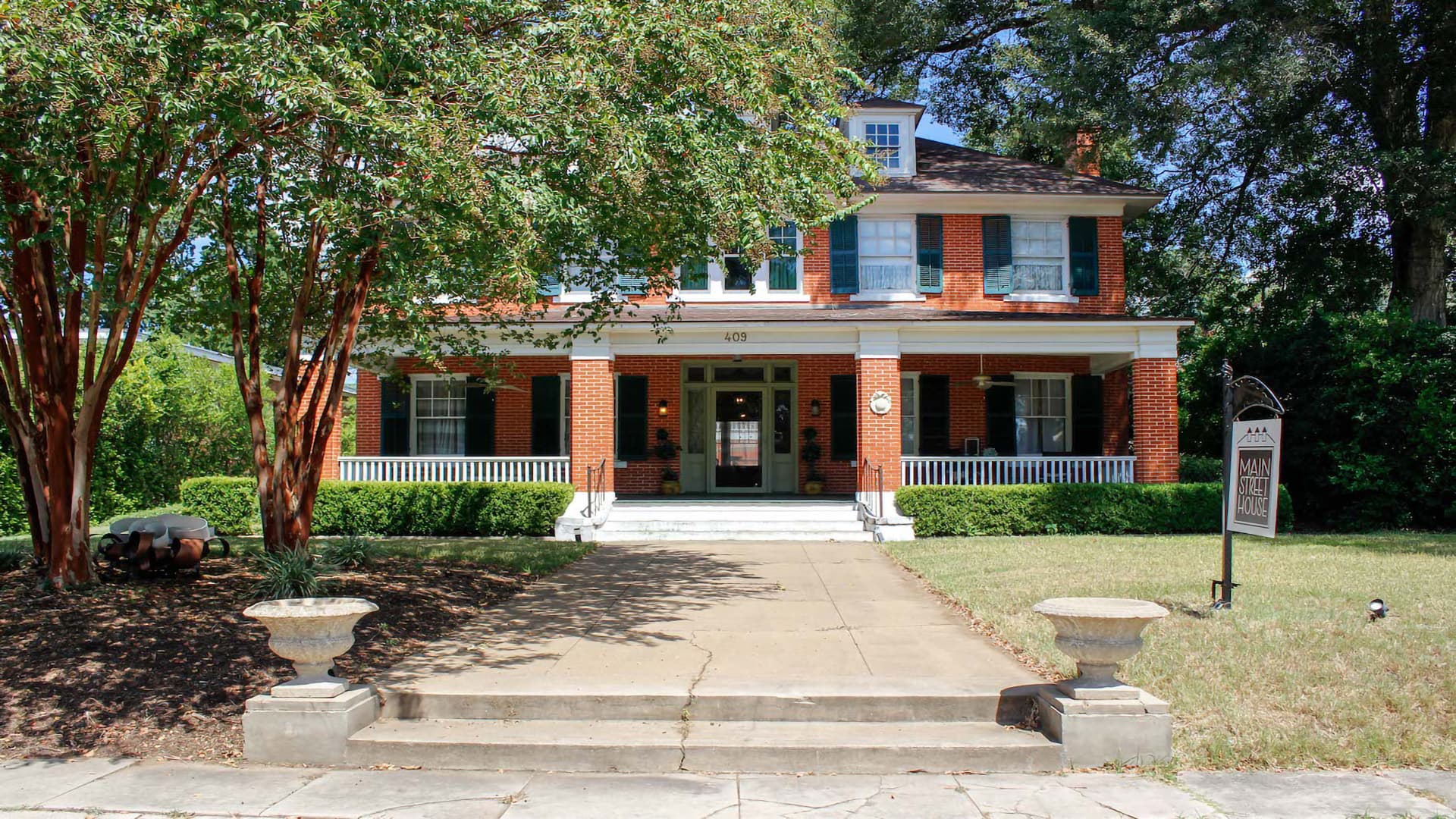 A historic brick house with a front porch, flanked by trees and a landscaped yard.